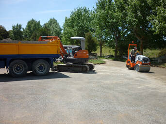 Roller working on levelling drive site after digger has unloaded gravel from truck, both also seen in photo